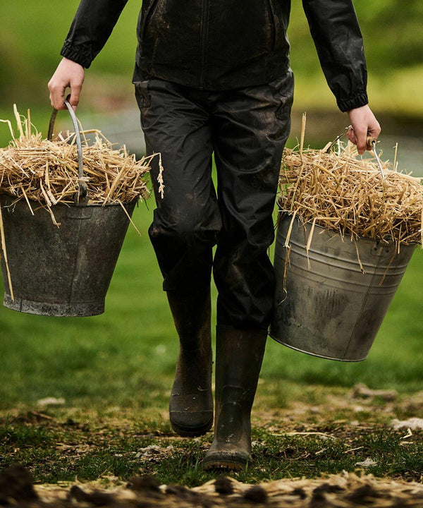Person carrying two RG254 buckets filled with straw while walking on grass, dressed in black Regatta rain gear and boots.