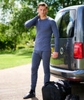 A person stands by a van, wearing a matching long-sleeve shirt and pants, sandals with socks, and resting their hand on their neck. A Regatta RG290 duffel bag sits on the ground beside them.
