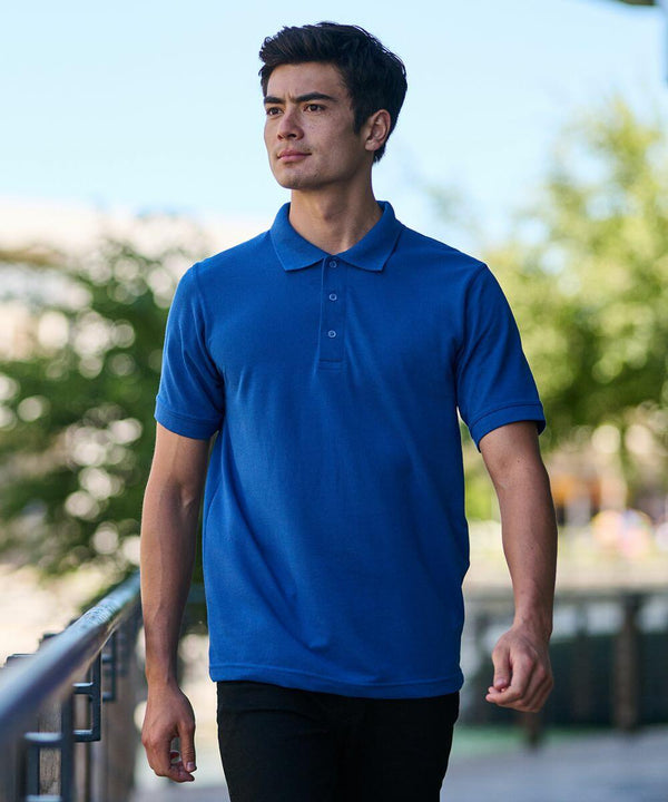 A person wearing a Regatta RG300 blue polo shirt and black pants is walking outdoors, with trees and buildings in the background.