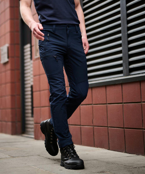 A person wearing dark blue cargo pants and black boots walks on a sidewalk next to a brick building with an RG307 Regatta metal vent.