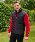 A man stands outdoors wearing a red RG340 sweater and a black padded vest, with his hands in his pockets. There is greenery in the background.
