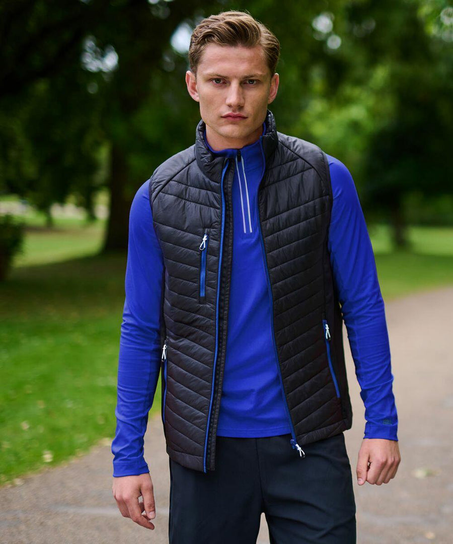A man wearing a blue long-sleeve shirt and a black Regatta RG341 vest stands on a pathway in a park with greenery in the background.