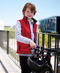 A boy with red hair wears a Regatta Junior Ablaze softshell bodywarmer over a grey long-sleeve shirt, standing on a bridge next to a black bike and holding a helmet. Modern buildings and trees can be seen in the background.