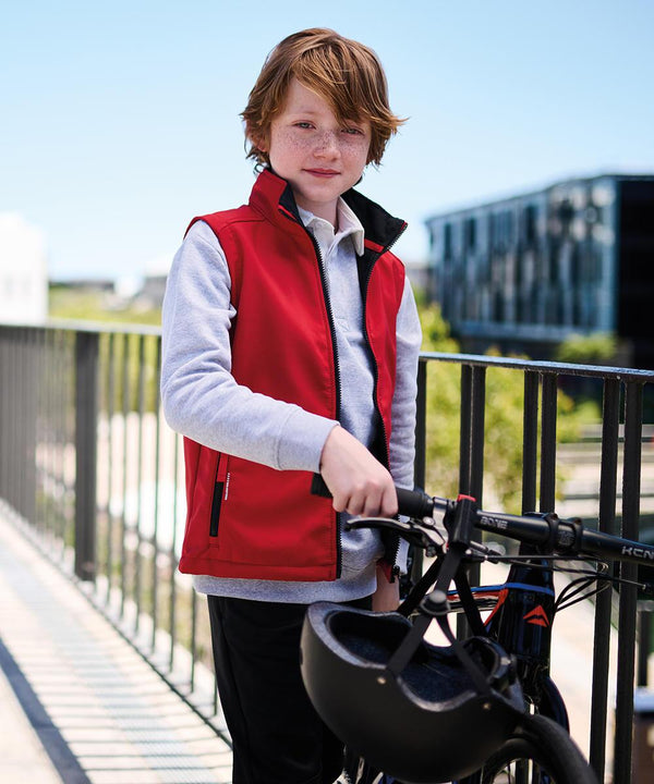 A boy with red hair wears a Regatta Junior Ablaze softshell bodywarmer over a grey long-sleeve shirt, standing on a bridge next to a black bike and holding a helmet. Modern buildings and trees can be seen in the background.