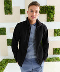 A man is standing indoors, wearing a black jacket over a striped shirt and blue pants. He is facing the camera with a neutral expression. The background features a modern design with green moss squares, while he holds an RG350 from Regatta.
