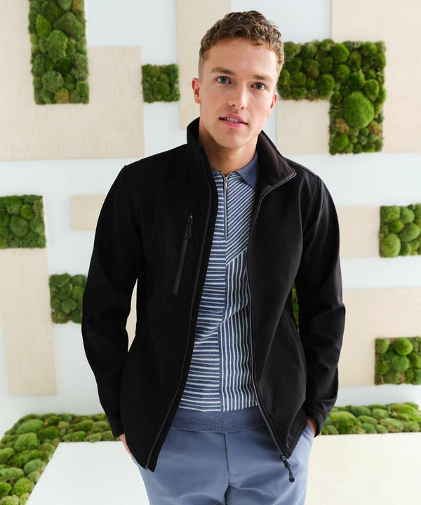 A man is standing indoors, wearing a black jacket over a striped shirt and blue pants. He is facing the camera with a neutral expression. The background features a modern design with green moss squares, while he holds an RG350 from Regatta.