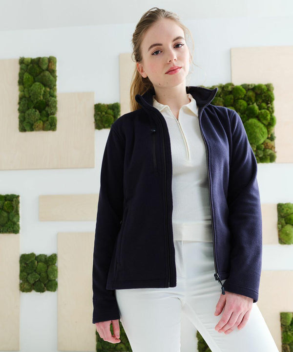 A woman in a Regatta RG361 dark jacket and white shirt stands in front of a wall with green moss decorations.