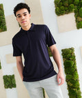 A young man wearing a black polo shirt and light grey pants stands in front of a wall decorated with green moss and wooden panels, showcasing the Regatta RG363.