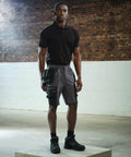 A person wearing a black RG366 polo shirt by Regatta, gray cargo shorts, and black boots stands in an industrial-style room with brick and white walls and concrete floor.