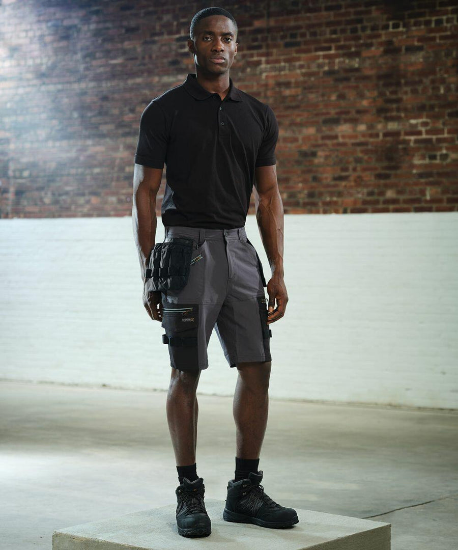 A person wearing a black RG366 polo shirt by Regatta, gray cargo shorts, and black boots stands in an industrial-style room with brick and white walls and concrete floor.