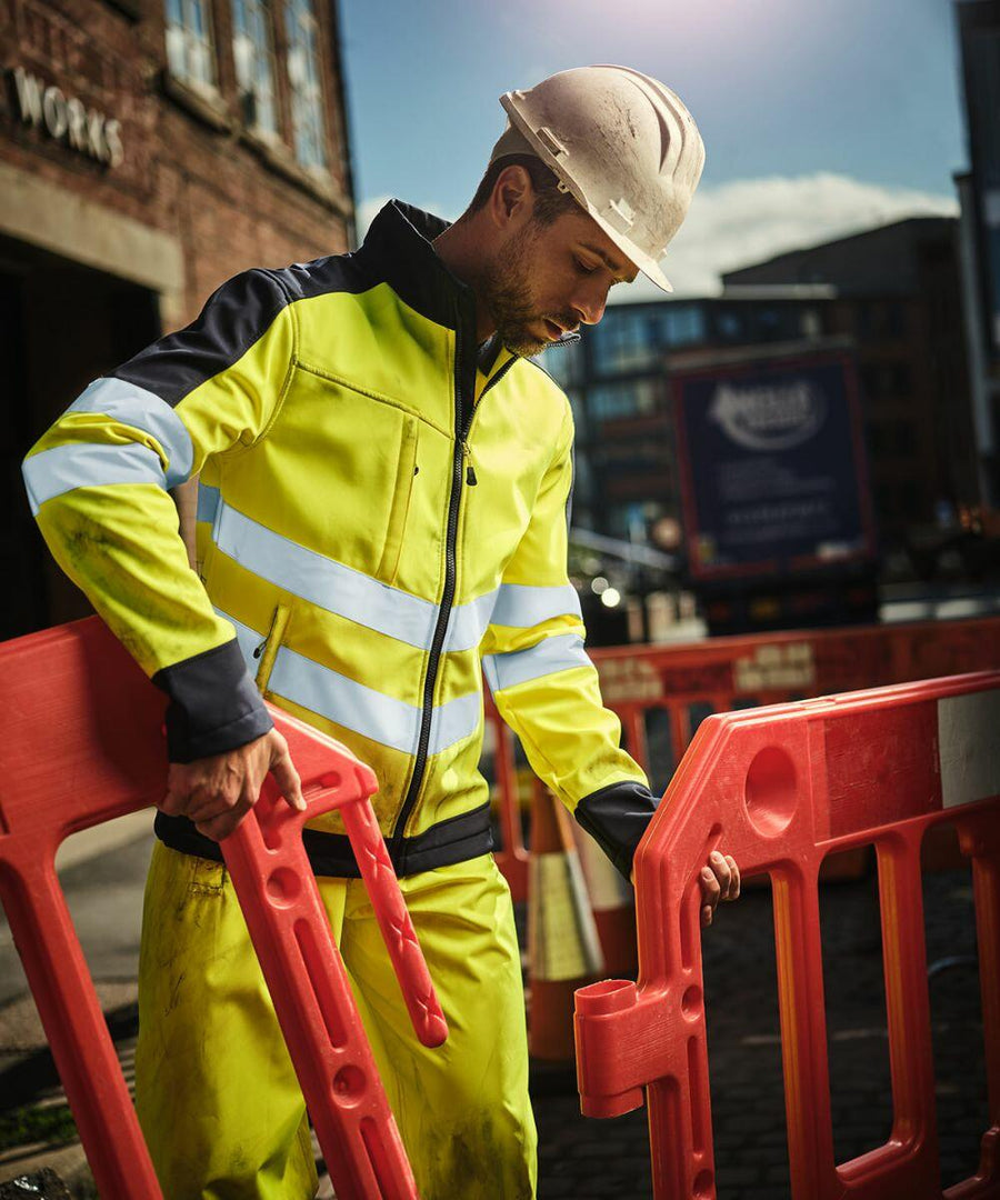 A worker in a yellow reflective jacket and hard hat adjusts RG382 safety barriers by Regatta at a construction site on a sunny day.