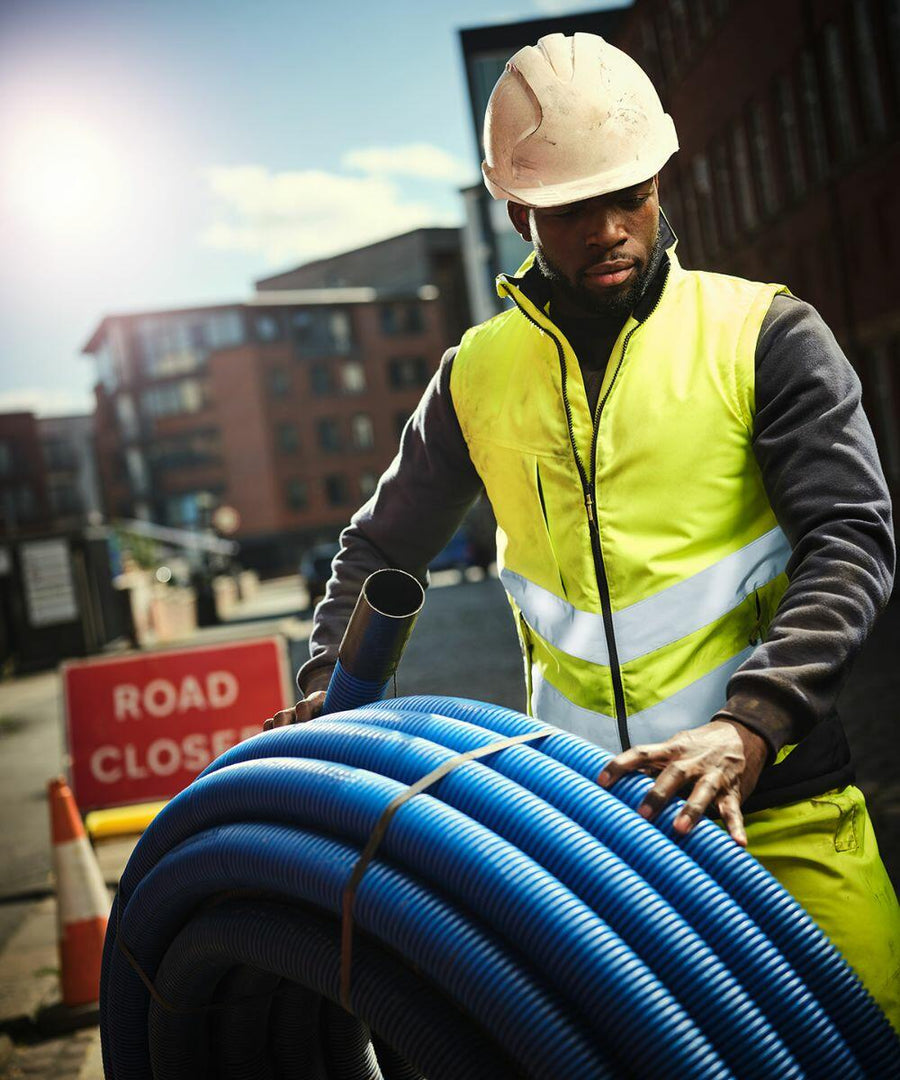 A construction worker wearing a high-visibility jacket and hard hat holds a coil of blue Regatta RG383 tubing on a street with a "Road Closed" sign in the background.