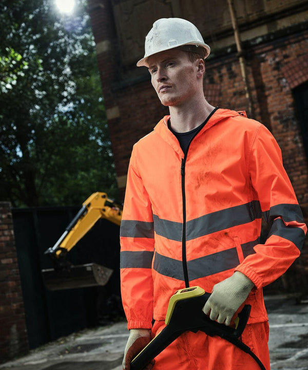 A worker in a Regatta RG385 high-visibility jacket and white hard hat holds equipment at a construction site with machinery and brick buildings in the background.