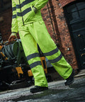 Person wearing high-visibility yellow rain gear with reflective stripes, standing on wet ground, and holding a Regatta RG386 garden hose reel in an industrial area.
