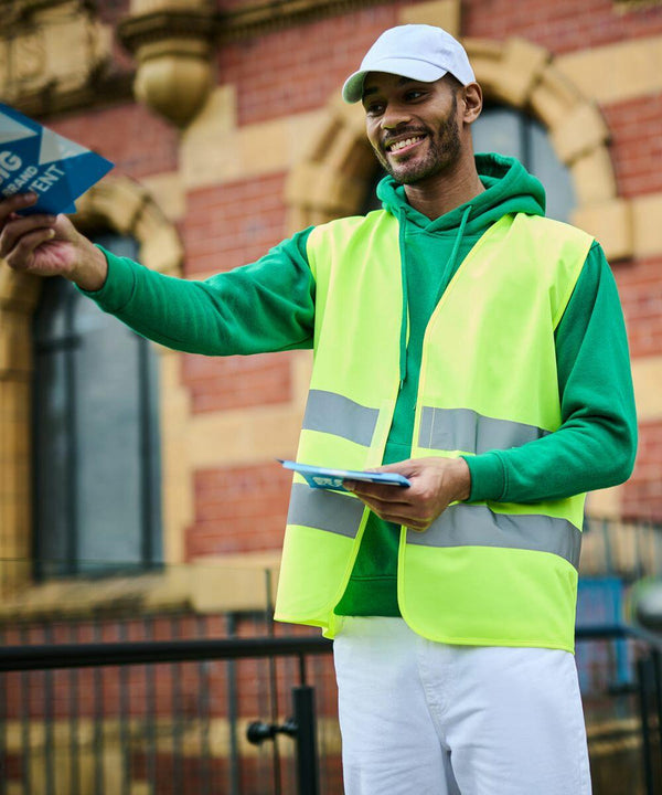 A person wearing a green RG471 hoodie by Regatta, a yellow safety vest, and a white cap is handing out flyers outdoors in front of a brick building.
