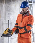 A construction worker in a Regatta High Visibility Hi-vis Pro contract Ablaze 2-layer softshell, blue hard hat, and gloves holds a yellow nail gun and tape measure at a site with concrete walls and metal fencing.