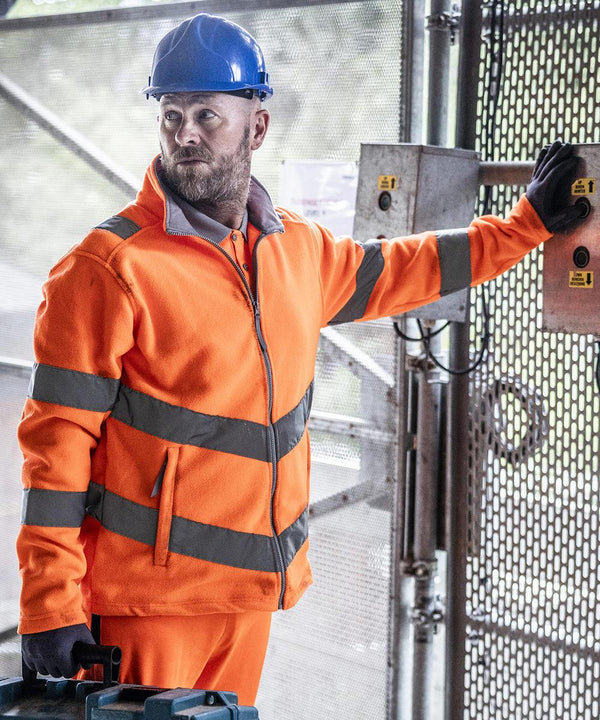 A man wearing a Regatta High Visibility Hi-vis Pro contract Thor fleece, a blue hard hat, and black gloves stands by a metal gate, pressing a control panel button with one hand and holding a blue tool case with the other.