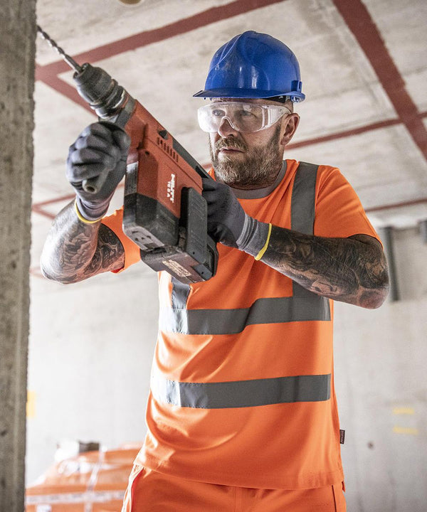 A construction worker wearing a Regatta High Visibility Hi-vis Pro contract t-shirt and a blue hard hat uses a power drill on a concrete wall inside a building under construction.