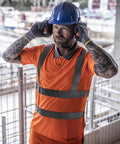 A construction worker with tattoos and a beard wears a Regatta High Visibility Hi-vis Pro contract polo, blue hard hat, gloves, and ear protection whilst standing at a worksite with metal fencing and equipment in the background.