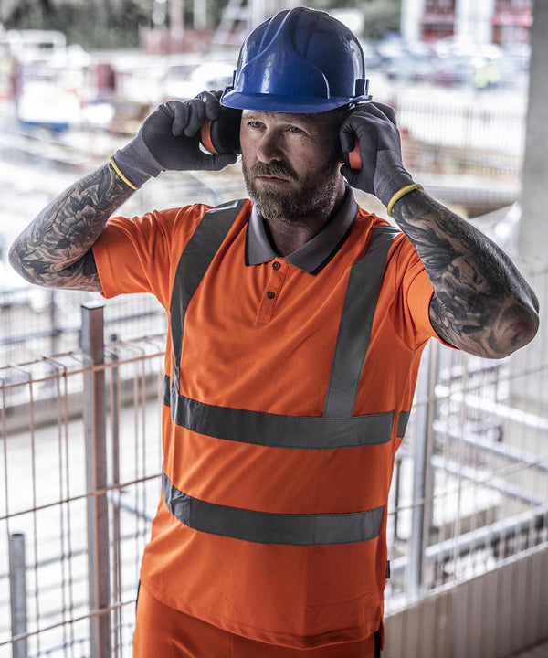 A construction worker with tattoos and a beard wears a Regatta High Visibility Hi-vis Pro contract polo, blue hard hat, gloves, and ear protection whilst standing at a worksite with metal fencing and equipment in the background.
