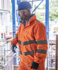 A man in a Regatta High Visibility Hi-vis Pro contract Dover jacket with reflective stripes, gloves, and a blue safety helmet stands sideways by a metal fence at a building site.