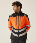 A man with curly hair models the Regatta High Visibility Hi-Vis X-Pro 3-layer softshell jacket (Class 2) in orange and black with reflective stripes, standing against a plain background with his hands behind his back, looking to his left.