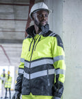 A man stands indoors at a construction site wearing a Regatta High Visibility Hi-Vis X-Pro Waterproof Shell Jacket (Class 2) and a white hard hat. In the background, two others in similar safety gear are visible.