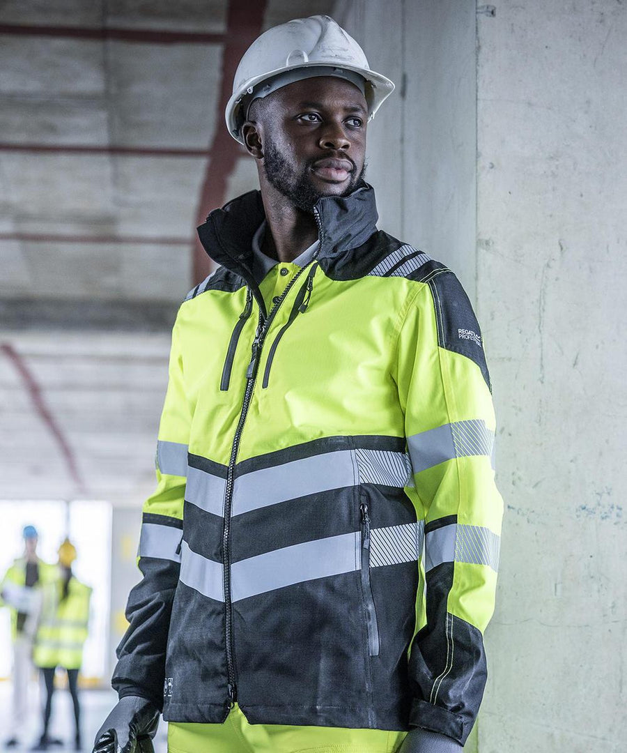A man stands indoors at a construction site wearing a Regatta High Visibility Hi-Vis X-Pro Waterproof Shell Jacket (Class 2) and a white hard hat. In the background, two others in similar safety gear are visible.