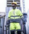 A construction worker in a Regatta High Visibility hi-vis two-tone thermal jacket and yellow safety helmet holds a tool, standing on a building site with building materials and a tall building in the background.