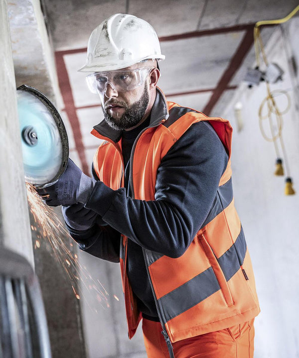 A construction worker wearing the Regatta High Visibility Hi-vis Pro contract Ablaze softshell bodywarmer, hard hat, and safety spectacles cuts through a concrete wall with a power saw as sparks fly in an unfinished interior with exposed wires.