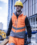 A construction worker in a Regatta High Visibility Hi-vis two-tone thermal bodywarmer carries wooden planks at an urban building site with tall buildings in the background.