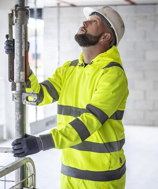 A man in a Regatta High Visibility Hi-Vis Pro contract hoodie, gloves, and a white hard hat inspects scaffolding inside a building with unfinished walls.
