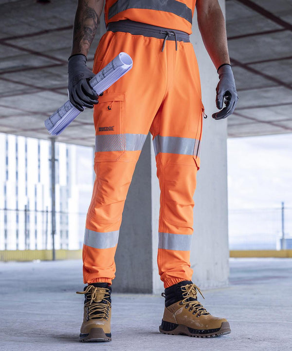 A person wearing Regatta High Visibility Hi-vis Prolite joggers and tan boots stands on a construction site, holding rolled blueprints and a mobile phone. Only the lower body is visible; the face and upper body are not shown.