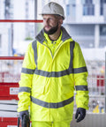 A man in a Regatta High Visibility Hi-vis Pro Contract Dover parka and a white hard hat stands at a building site, looking to the side.