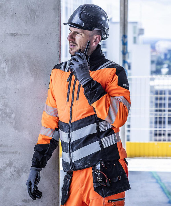 A construction worker in a Regatta High Visibility Hi-vis X-pro hybrid jacket (Class 2), gloves, and a black hard hat stands by a concrete wall, speaking into a two-way radio at a building site.