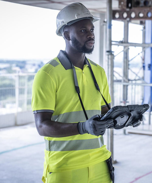 A man in a Regatta High Visibility Hi-vis pro comfort polycotton polo, white hard hat, and gloves holds a tablet at a construction site with scaffolding and railings in the background.