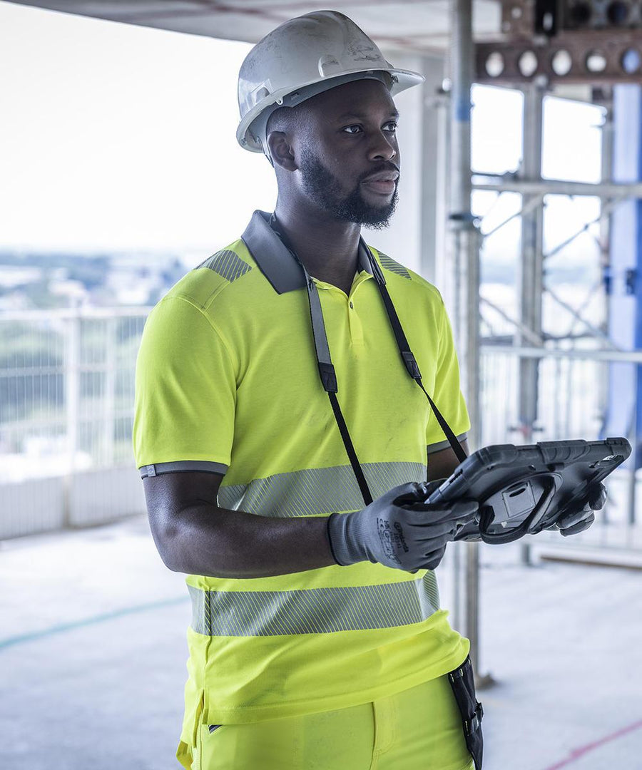 A man in a Regatta High Visibility Hi-vis pro comfort polycotton polo, white hard hat, and gloves holds a tablet at a construction site with scaffolding and railings in the background.
