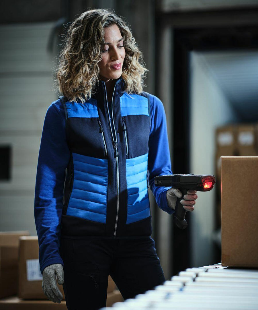 A woman in a blue and black jacket scans a cardboard box with a Regatta RG542 handheld barcode scanner in a warehouse.
