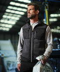 A man with light brown hair and a beard is wearing a black vest over a long-sleeved white shirt, holding a white Regatta RG544 bag in an industrial setting with overhead lights.