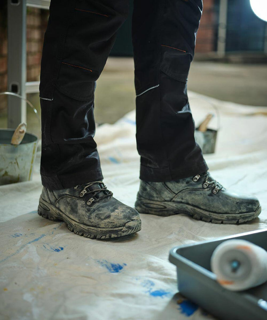 A person wearing black work pants and dusty black boots stands on a white tarp near a gray paint tray and various painting supplies, including the Regatta RG565. The scene suggests an ongoing painting or renovation project.