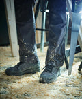 Person's legs in Regatta RG567 work pants covered with sawdust, holding a hand saw, standing on a sawdust-covered floor in a workshop.