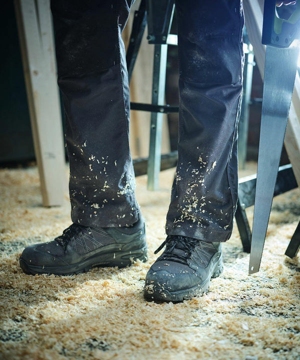 Person's legs in Regatta RG567 work pants covered with sawdust, holding a hand saw, standing on a sawdust-covered floor in a workshop.