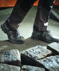 A person wearing dark work pants and black work boots stands on a tiled surface next to several cobblestones, with Regatta's RG568 nearby.