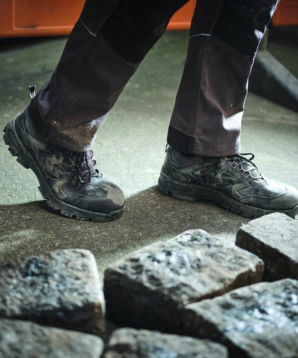 A person wearing dark work pants and black work boots stands on a tiled surface next to several cobblestones, with Regatta's RG568 nearby.