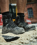 Close-up of a person wearing Regatta RG575 dusty black work boots standing on sandy ground at a construction site, with blurred machinery and a brick building in the background.