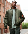 A man wearing a Regatta RG588 olive green bomber jacket and green pants stands in an urban alley with brick buildings.