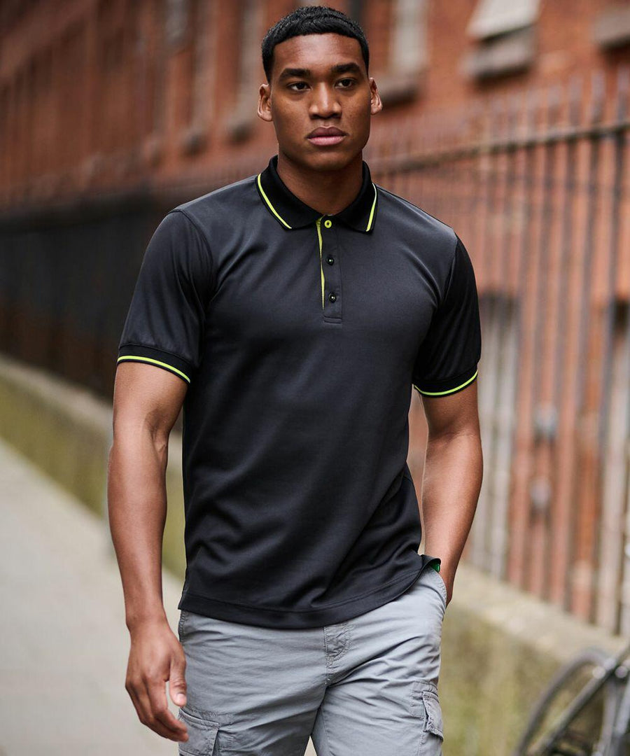 A man in a Regatta RG596 black polo shirt with neon green accents and gray shorts walks down a city street near a red brick building and iron fence.