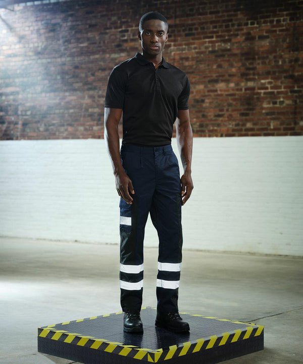 A man stands on a black and yellow RG609 platform by Regatta, wearing a black polo shirt and dark pants with reflective stripes, in a room with brick and white walls.