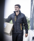 A man wears the Regatta Professional Pro universal jacket with orange zips as he carries a cardboard box outside near a building with glass windows.