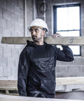 A person in a Regatta Professional Pro trade hoodie and white hard hat carries a wooden beam inside a partially built structure with exposed brick walls and a window behind them.